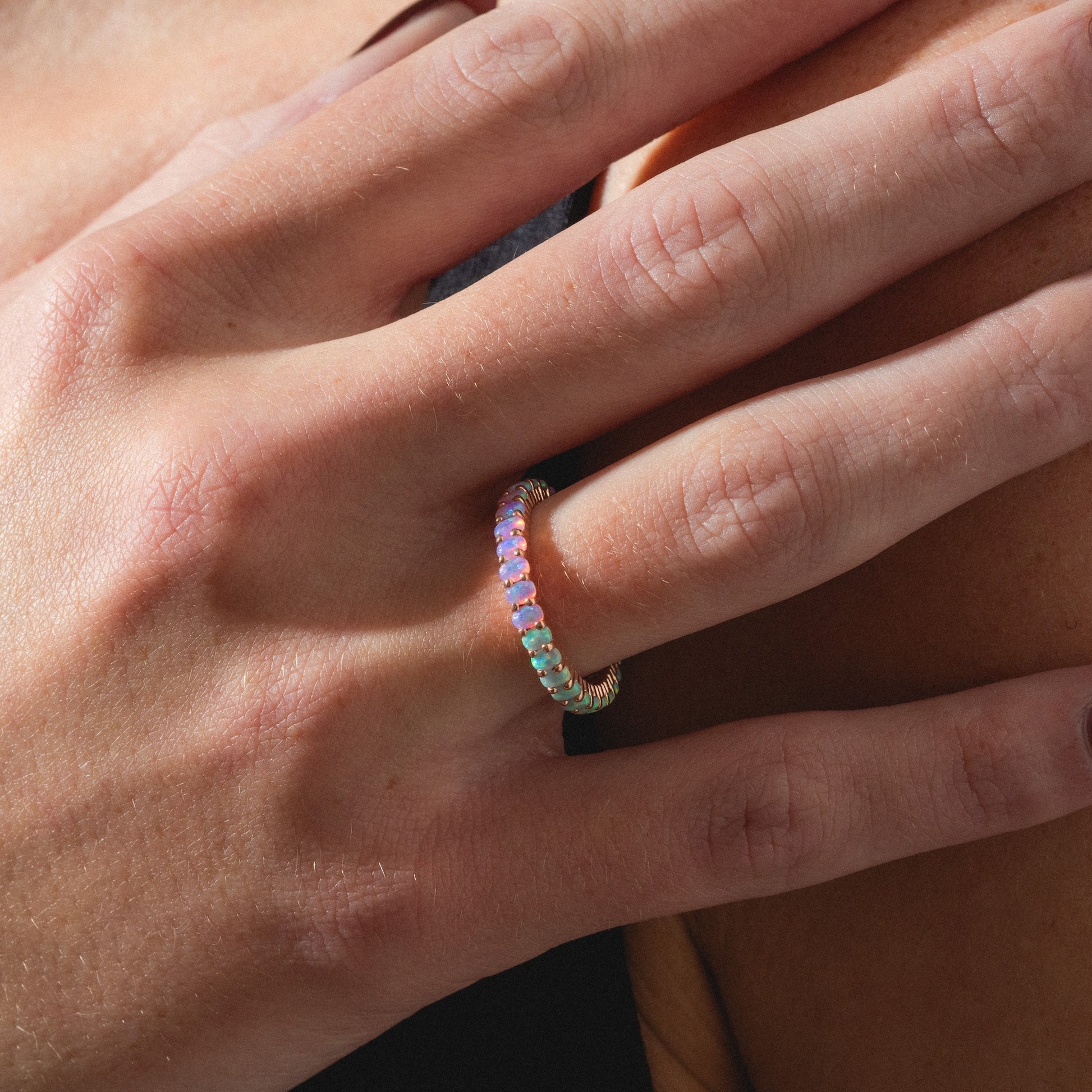 A close-up of a hand wearing the Opal Ombre Dual Band, a gold ring set with small round multicolored opal stones, gently resting on skin as soft natural light highlights its vibrant hues.
