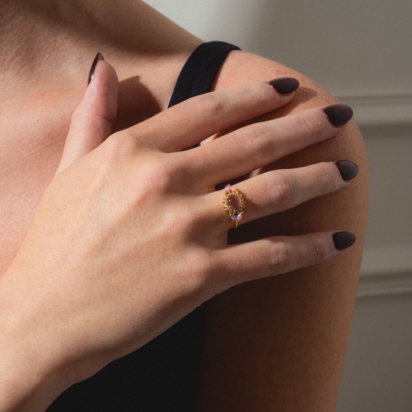 A person in a black strap top rests their hand on their shoulder, displaying the Pink Opal and Cats Eye Moonstone Sunflower Ring with dark polished nails.