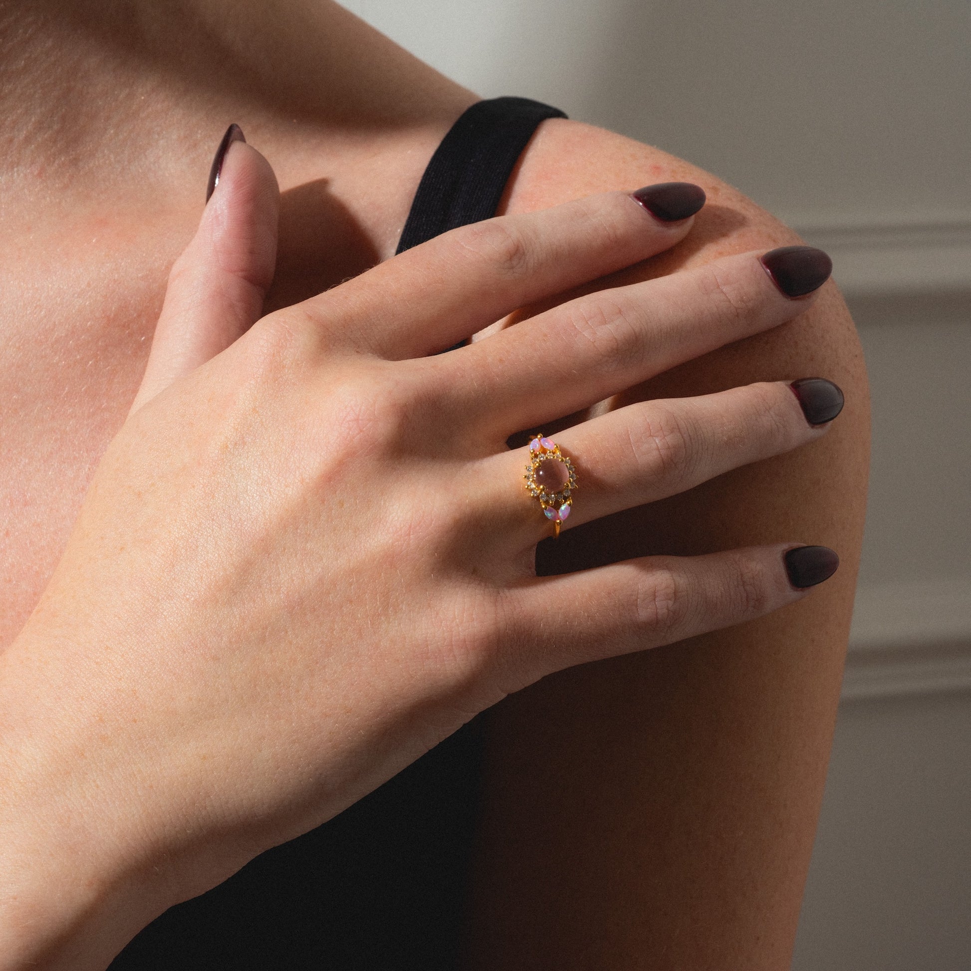 A person in a black strap top rests their hand on their shoulder, displaying the Pink Opal and Cats Eye Moonstone Sunflower Ring with dark polished nails.