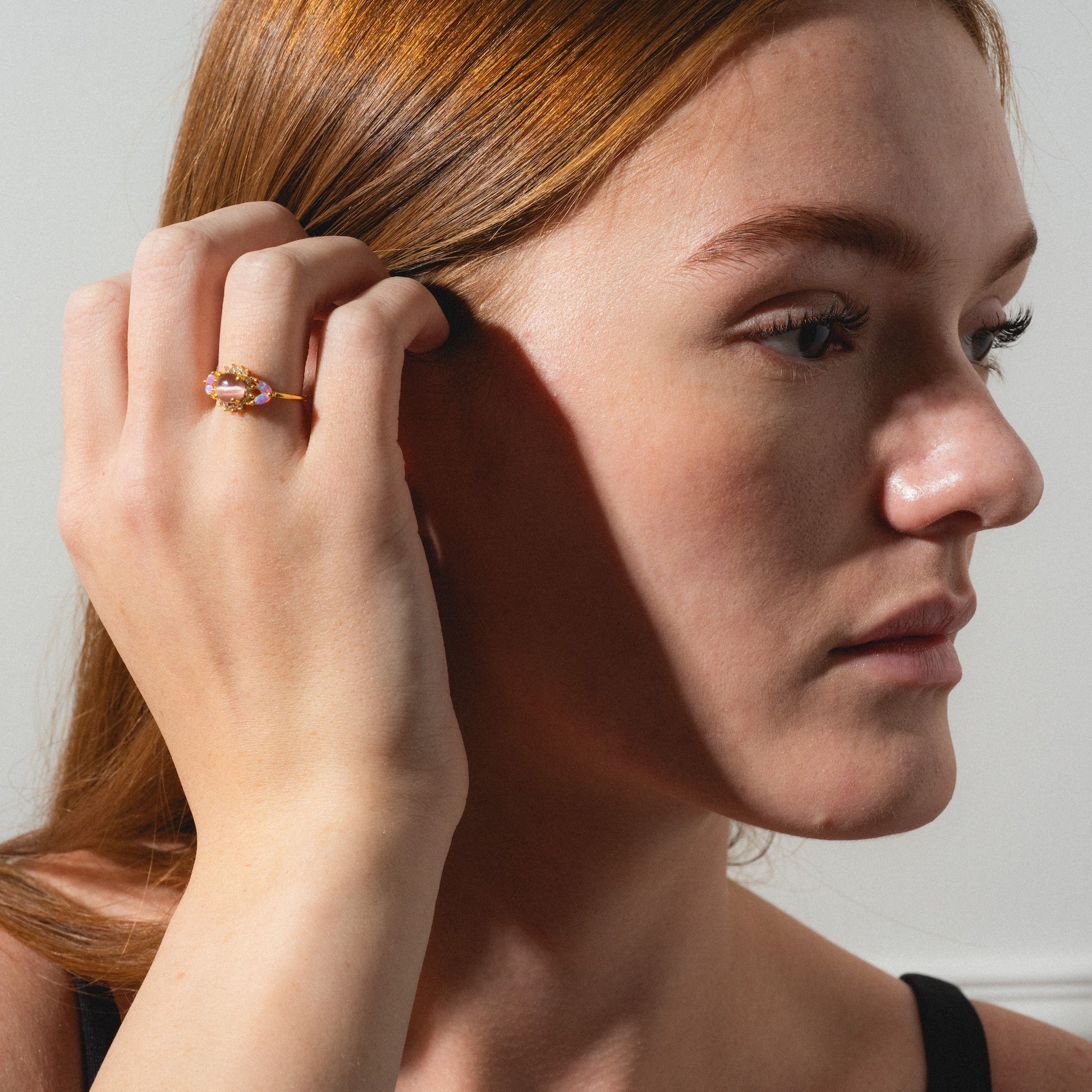 A woman with long red hair gently touches her ear, wearing the Pink Opal and Cats Eye Moonstone Sunflower Ring on her finger. She looks to the side as soft natural light highlights her face.