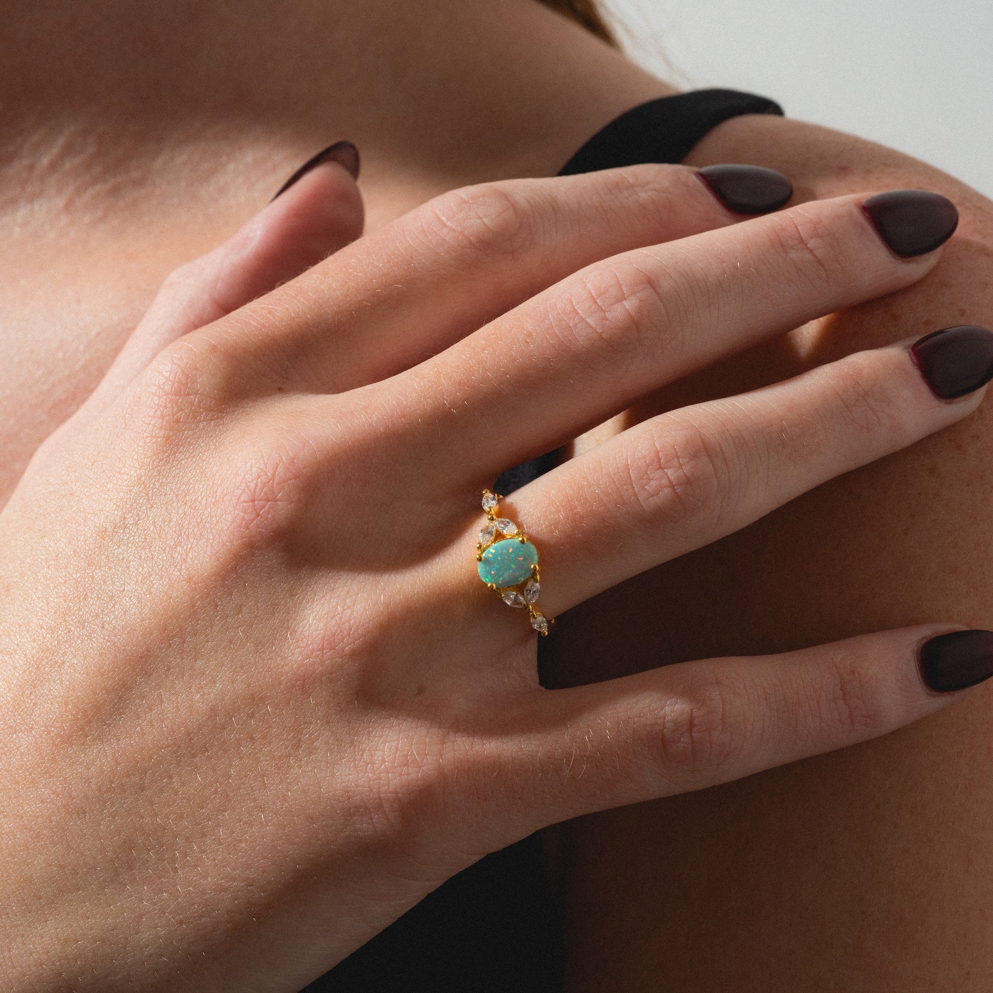 A hand with dark burgundy nails wears the Opal Enchanted Ring, a gold piece featuring a large oval opal surrounded by small gems. The person is dressed in a black strap garment.