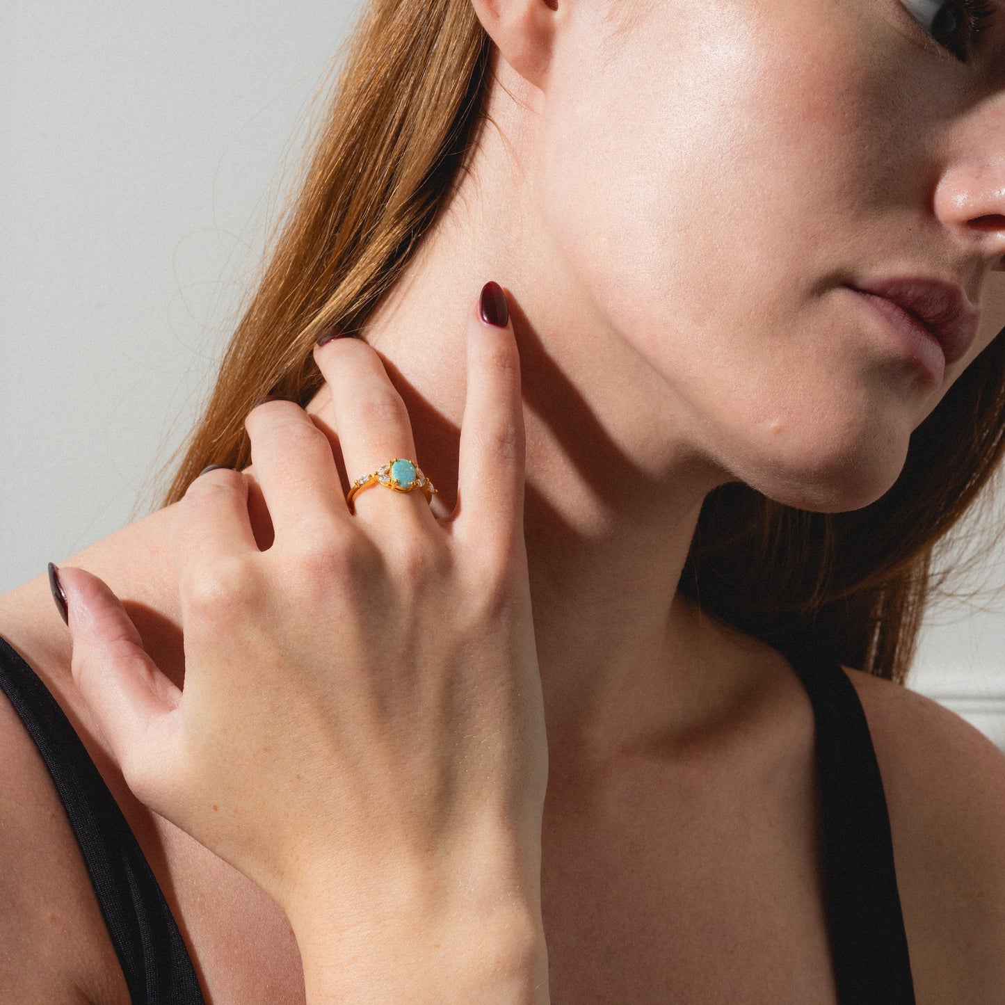 A woman with fair skin and long brown hair, wearing dark red nail polish and a black top, touches her neck with her left hand while showcasing the Opal Enchanted Ring.