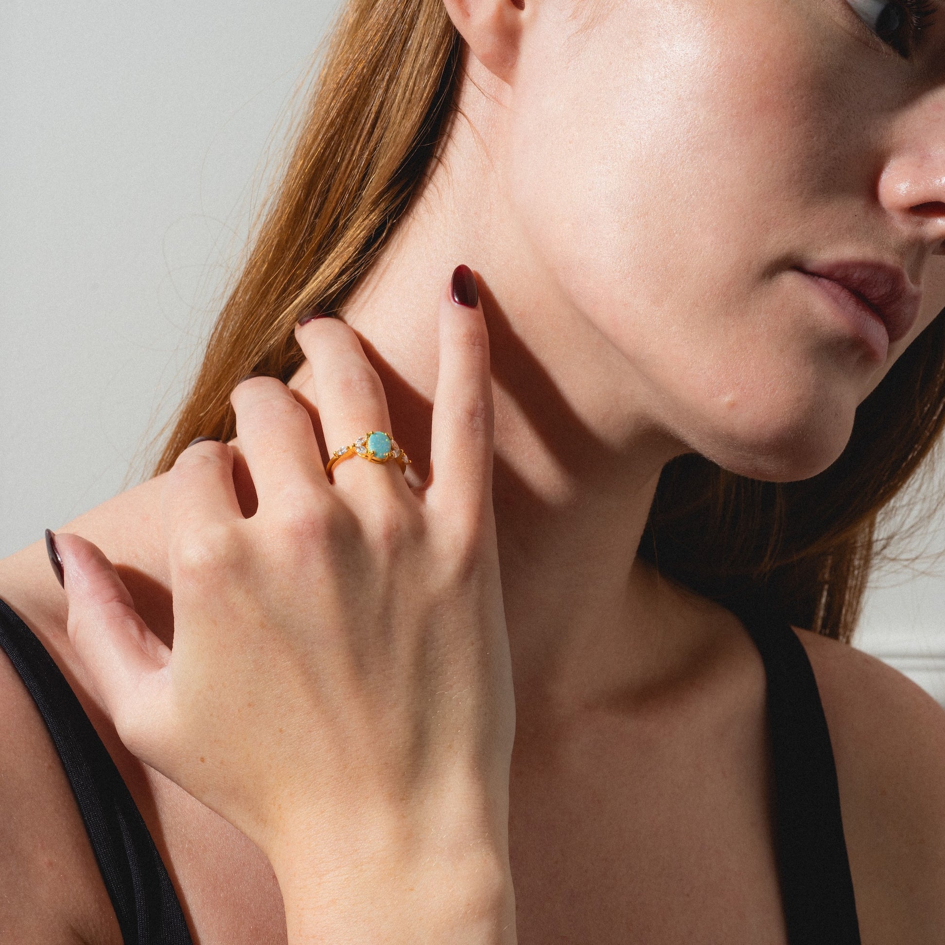 A woman with fair skin and long brown hair, wearing dark red nail polish and a black top, touches her neck with her left hand while showcasing the Opal Enchanted Ring.