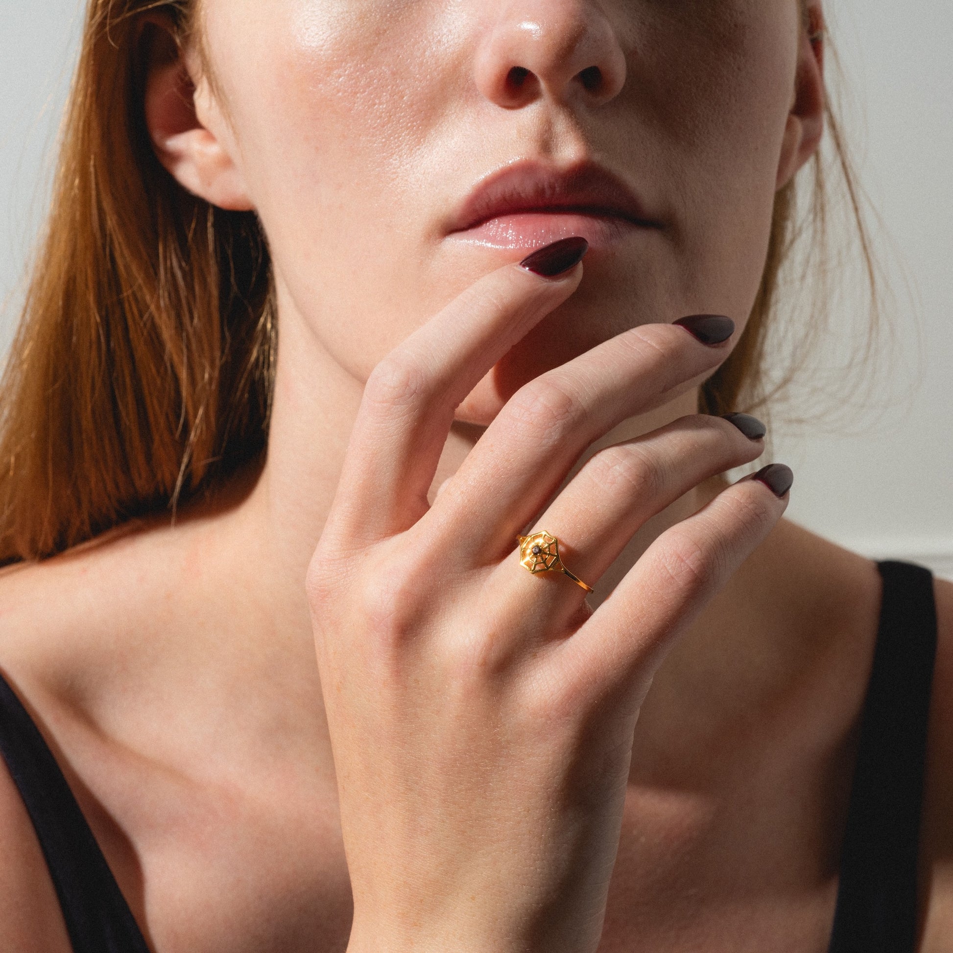 A woman with light skin and long hair touches her chin, showcasing the Limited Edition Black Widow Garnet Spiderweb Ring on her finger. She wears dark nail polish, a black top, and her face is partially visible.