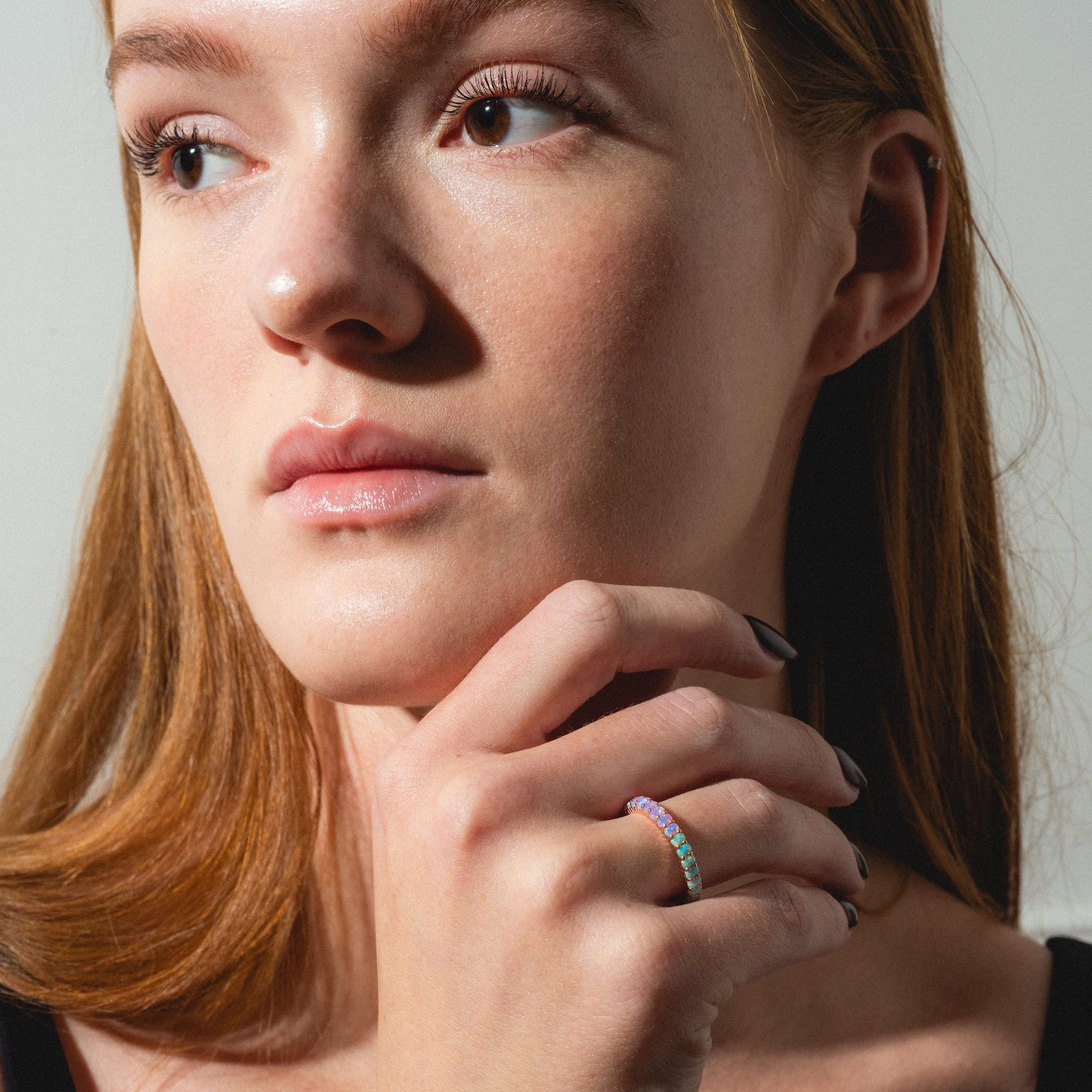 A woman with fair skin and straight auburn hair looks to the side, touching her face as she wears the Opal Ombre Dual Band—a colorful gemstone ring. She has minimal makeup and a black top.