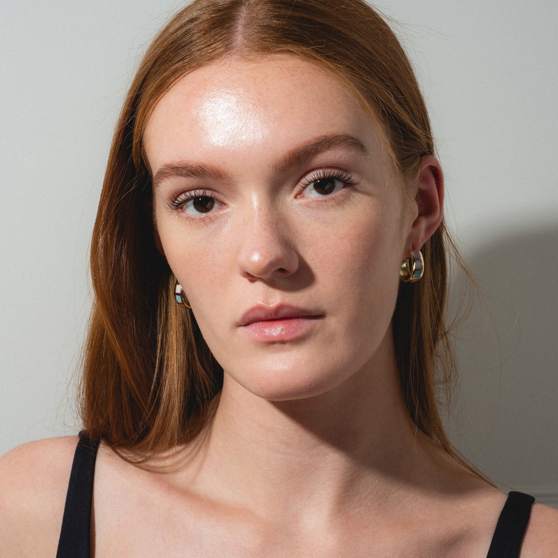 A young woman with long red hair wears Opal Ocean Lover Hoops and a black top, looking directly at the camera with a neutral expression against a plain light background.