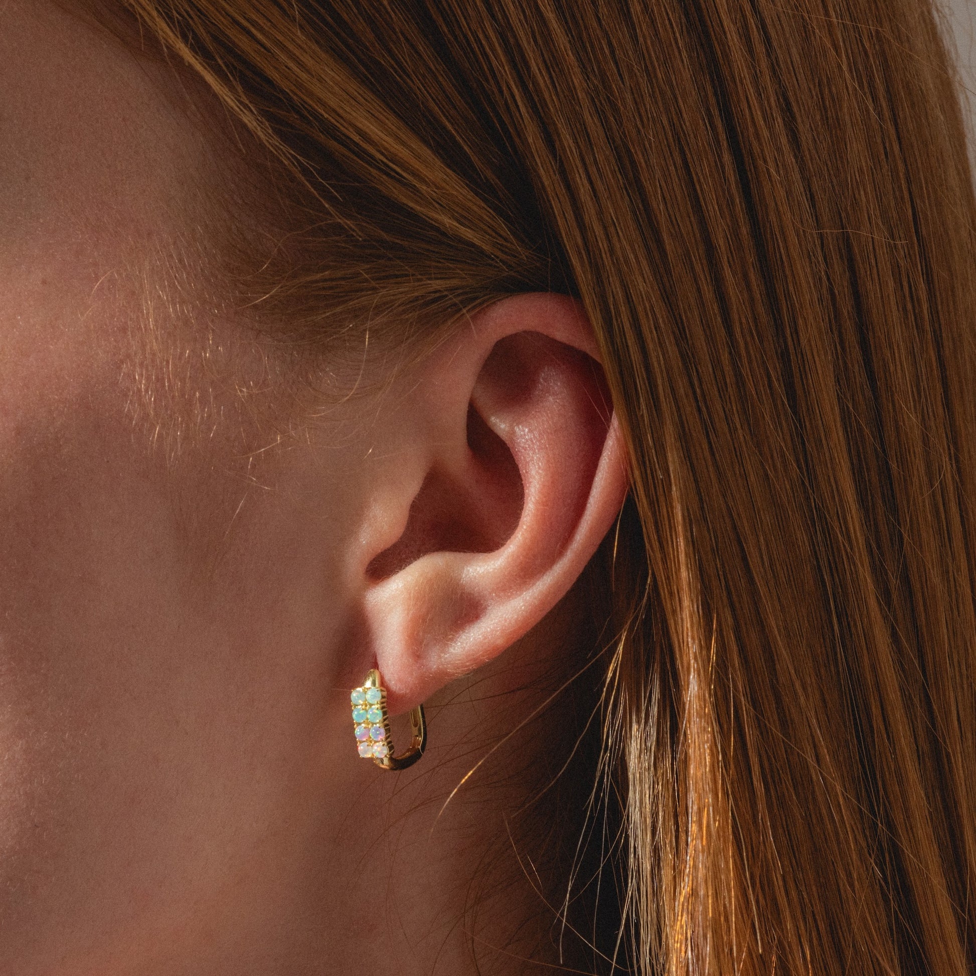Close-up of a person with straight reddish-brown hair wearing Opal Ombre Rue Huggies—small gold hoops embellished with green and white gemstones. Soft lighting highlights the ear and jewelry details.