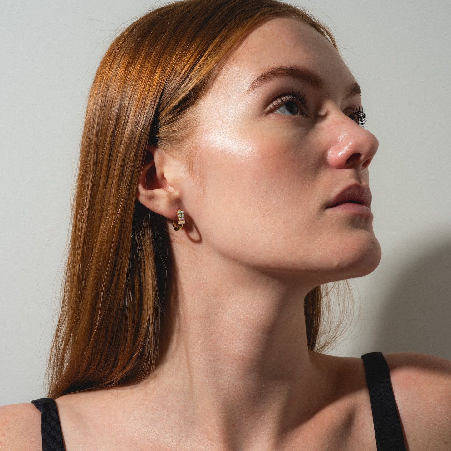 A woman with long straight reddish-brown hair and fair skin looks up to the right, wearing a black top and Opal Ombre Rue Huggies. The lighting highlights her glowing complexion against a plain background.