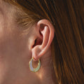 A close-up of a person's ear with straight auburn hair, wearing Opal Ombre Lunar Hoops—circular gold earrings featuring a pearly, iridescent inlay.