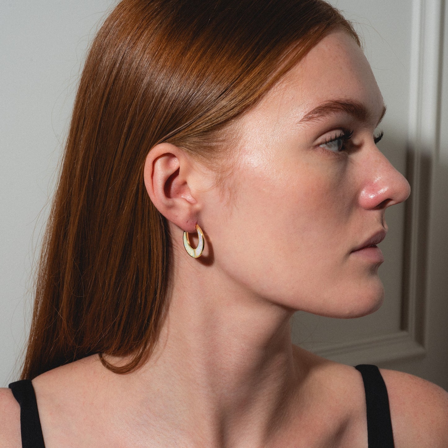 A woman with long, straight auburn hair and fair skin is shown in profile, wearing Opal Ombre Lunar Hoops and a black top. She stands against a white wall, soft lighting highlighting her features.