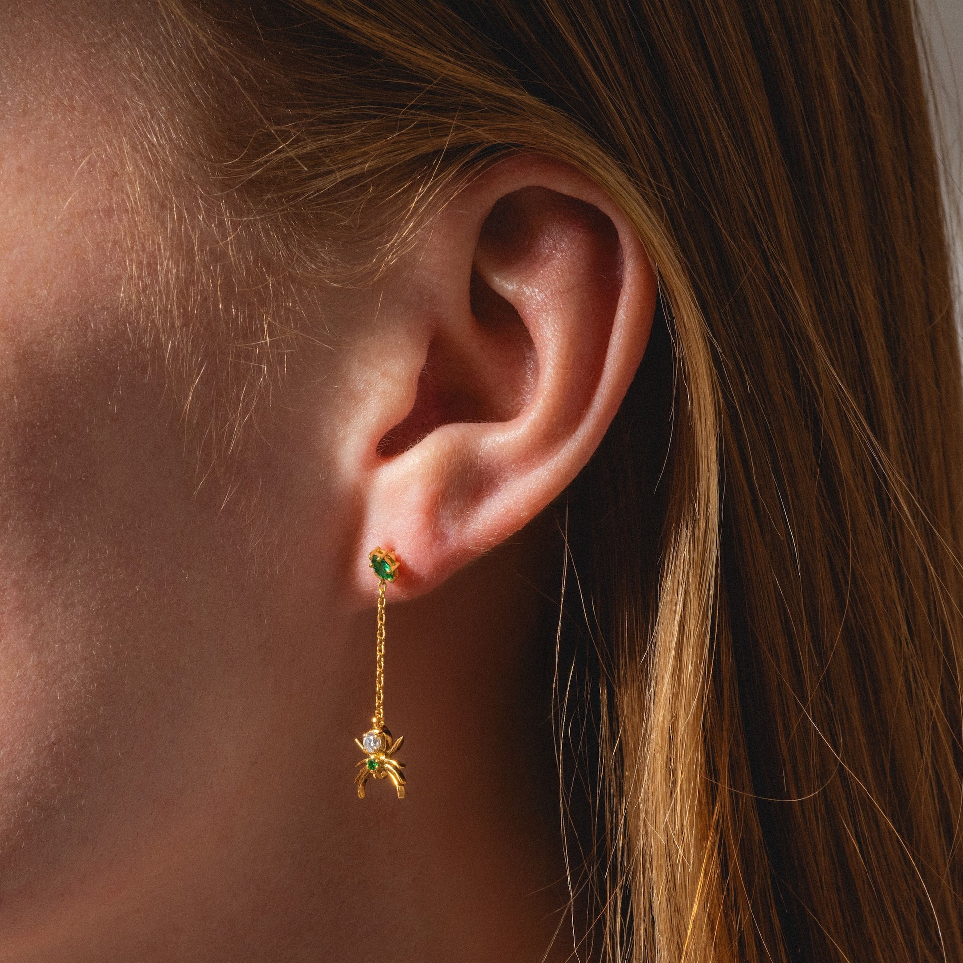 A close-up shows a person with straight, light brown hair tucked behind their ear, wearing the Limited Edition Tangled Webs Spider Drop Earrings—gold with a green gemstone stud and a dangling spider-shaped charm.