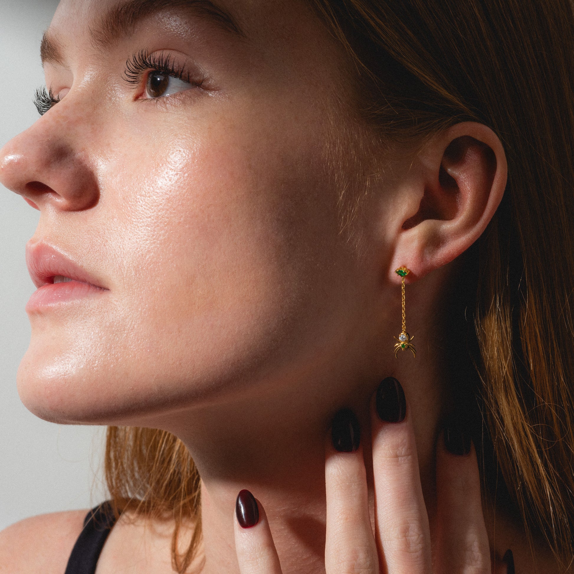 A light-skinned woman with auburn hair looks left, wearing Limited Edition Tangled Webs Spider Drop Earrings—gold with a small green gem. Her dark-polished nails gently touch her neck near the earring.