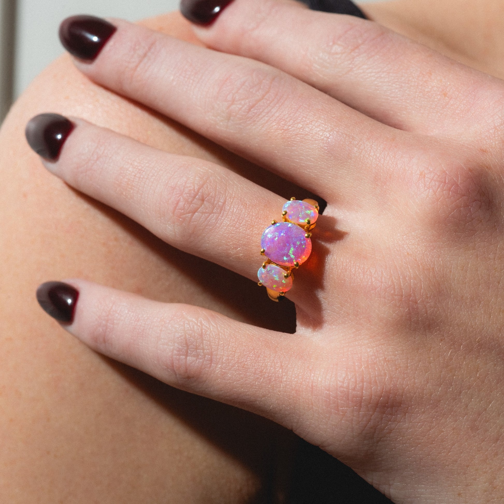 A hand with dark, polished nails wears the Pink Opal Gum Drop Ring—a gold ring set with three oval pink opal gemstones showing iridescent colors—against a neutral background.