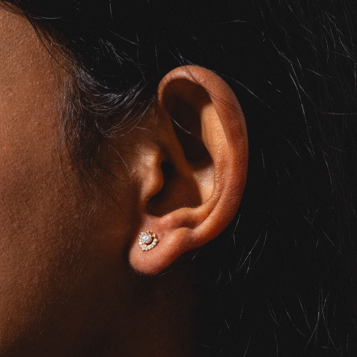 Close-up of a person's ear wearing Tosia Diamond Studs, small round gold earrings with a central stone encircled by tiny clear stones, shown against a dark background.