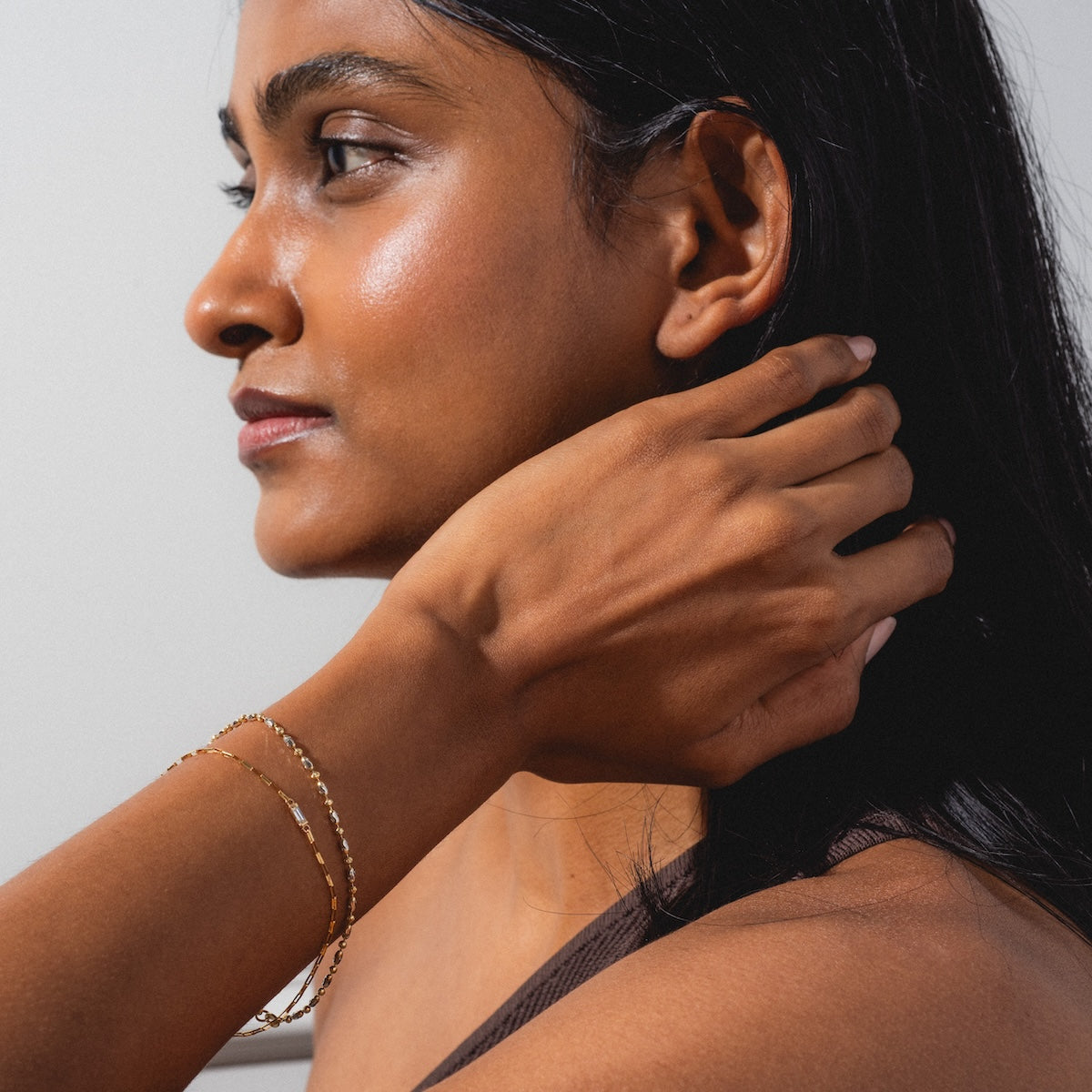 A woman with smooth dark skin and long black hair touches her neck, wearing two Sunblink Diamond Bracelets. She gazes to the side in soft natural light against a plain background.
