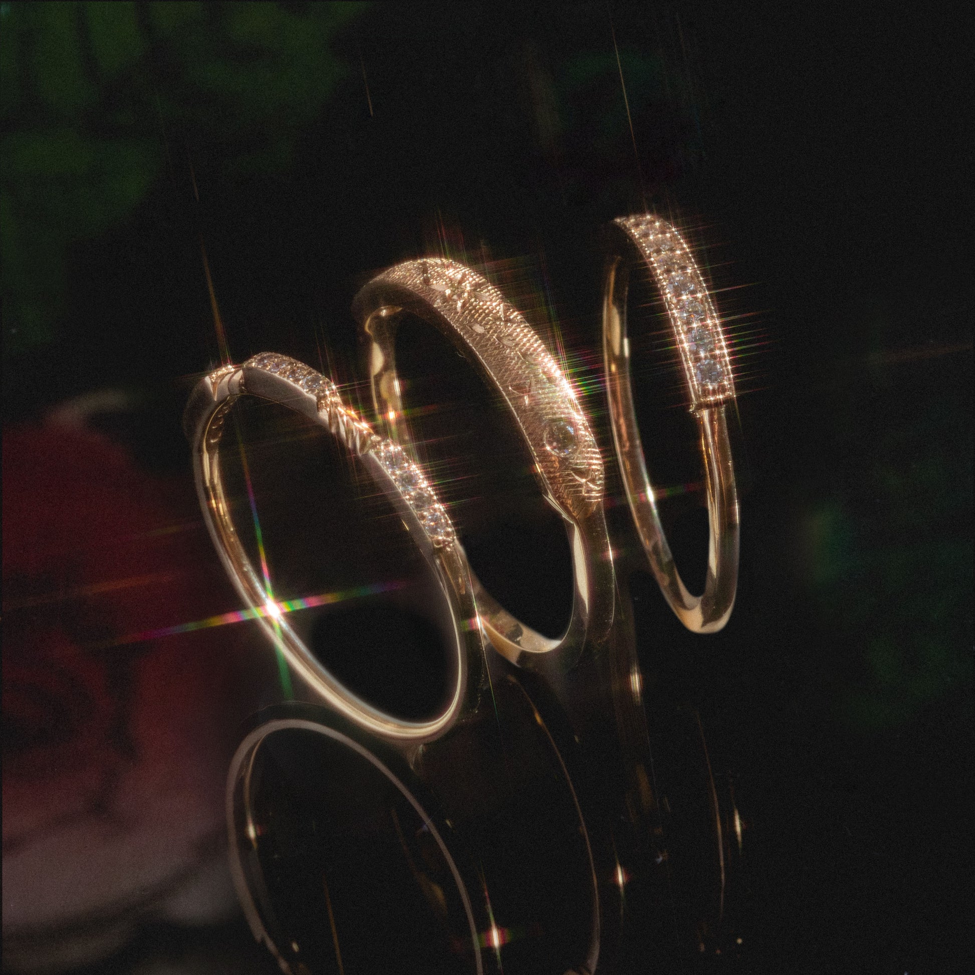 Three Clyde Rings with intricate designs and sparkling gemstones are displayed on a reflective black surface, illuminated by soft light that creates a starburst effect across the jewelry.