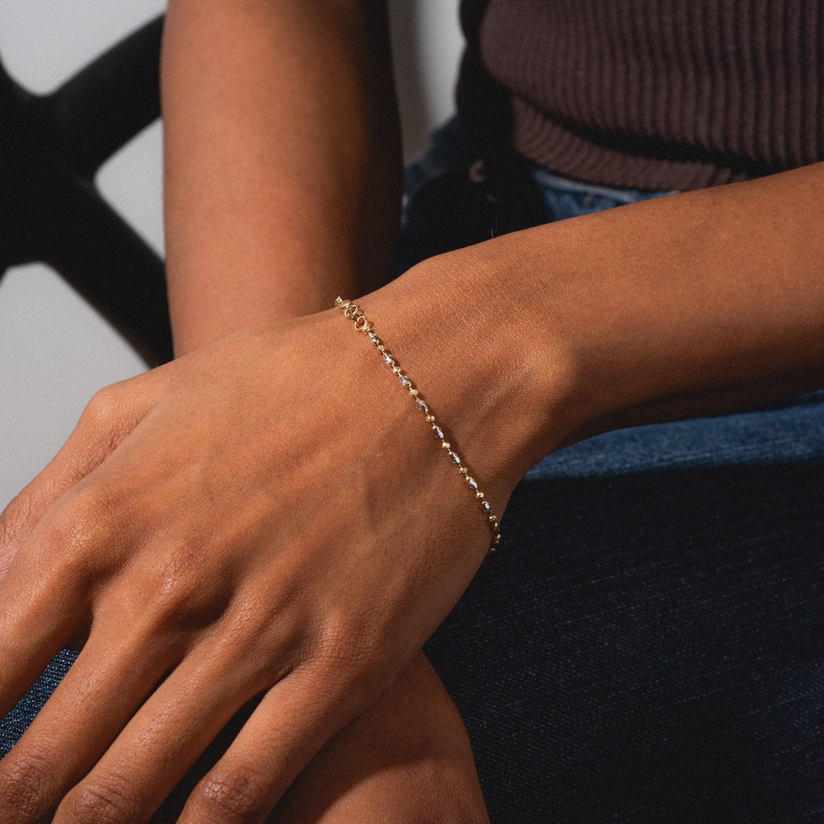 A close-up of a hand and wrist wearing the Bubble Two Tone Bracelet. The person is dressed in dark blue jeans and a sleeveless brown top, with a softly blurred background.