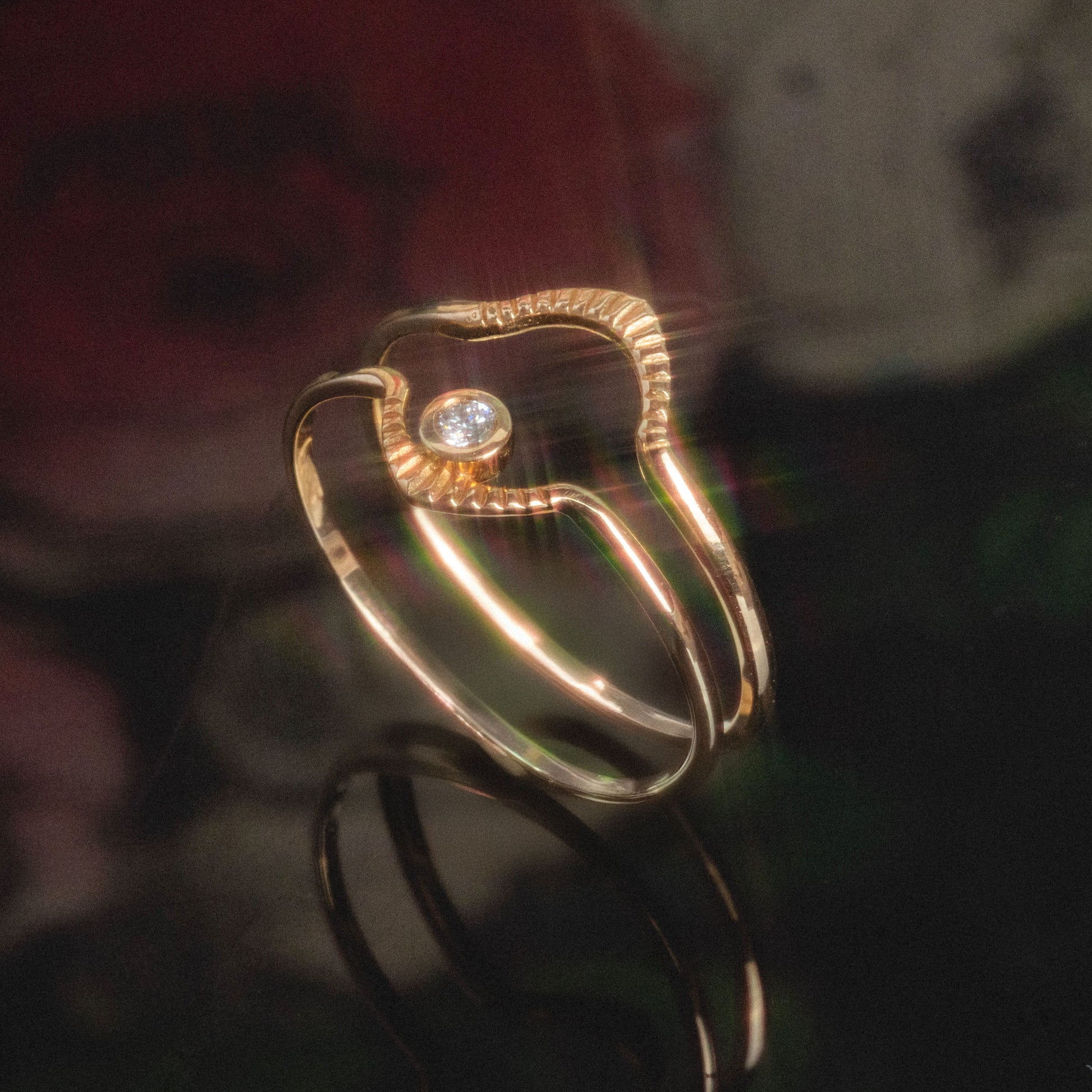 A close-up of the Corrie Ring, featuring a unique wavy gold design with a single round diamond at its center, set against a dark, softly blurred background with hints of red and green.