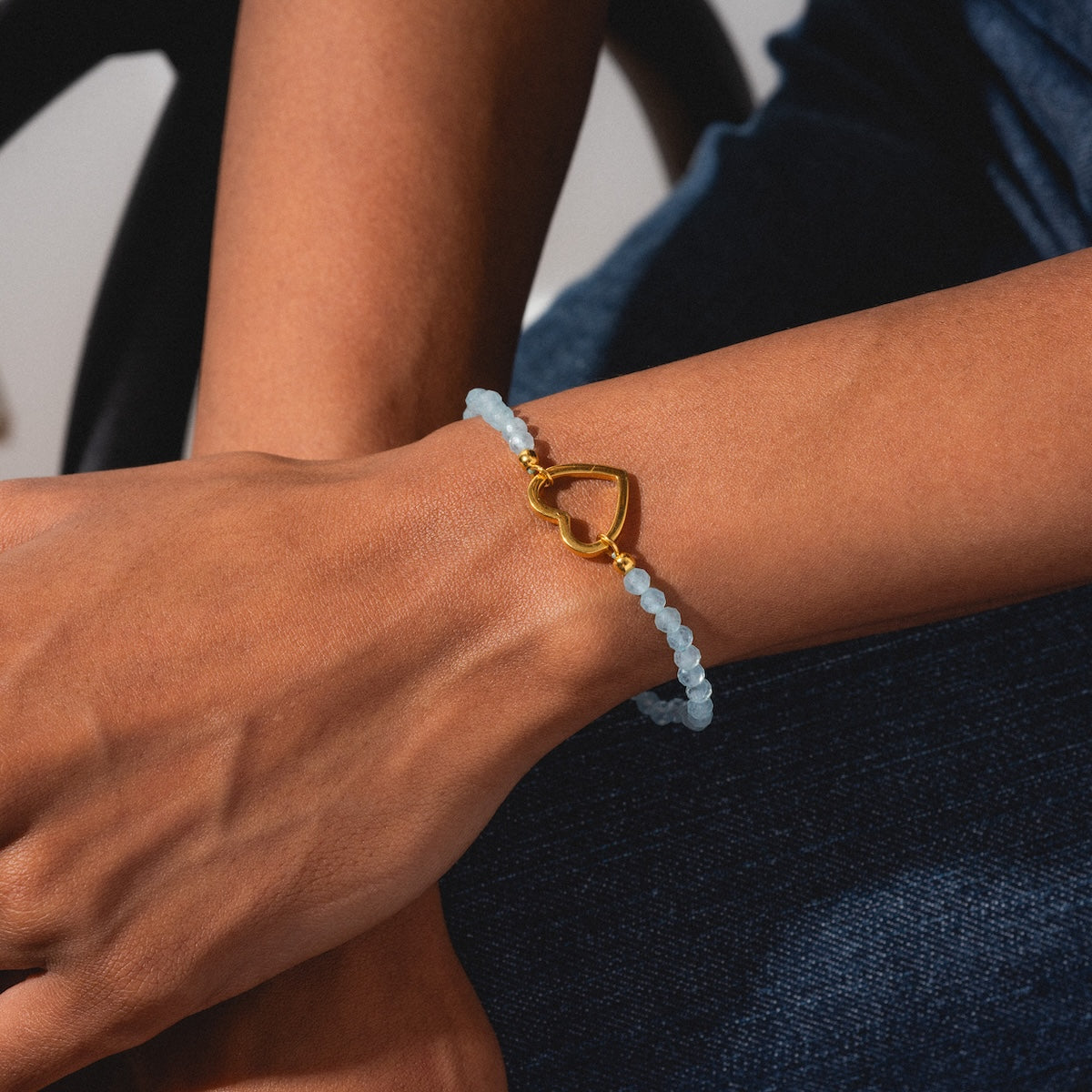 A close-up of a wrist wearing the Aquamarine Beaded Heart Clasp Bracelet, featuring light blue beads and a gold heart-shaped charm, with the hand resting on a lap in dark blue jeans.