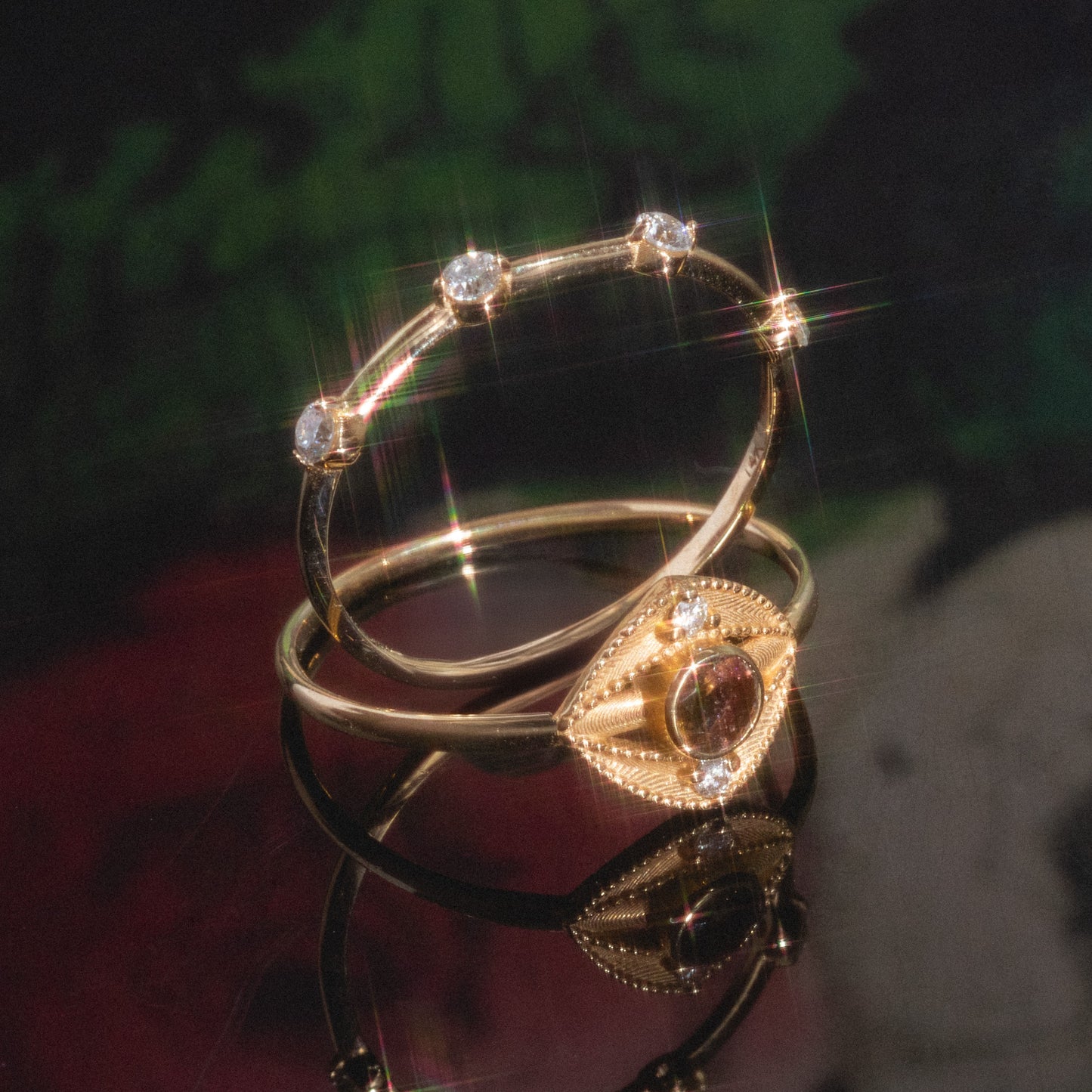 The Munro Ring rests on a reflective surface; it showcases an eye-shaped design with a central gemstone and small diamonds along the band, set against a softly blurred background.