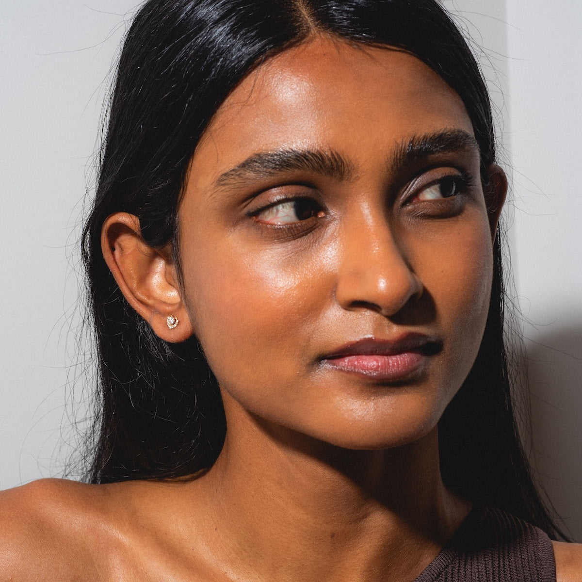A woman with long dark hair and glowing skin smiles gently as she looks to the side, wearing a brown off-the-shoulder top and Tosia Diamond Studs against a plain light background.