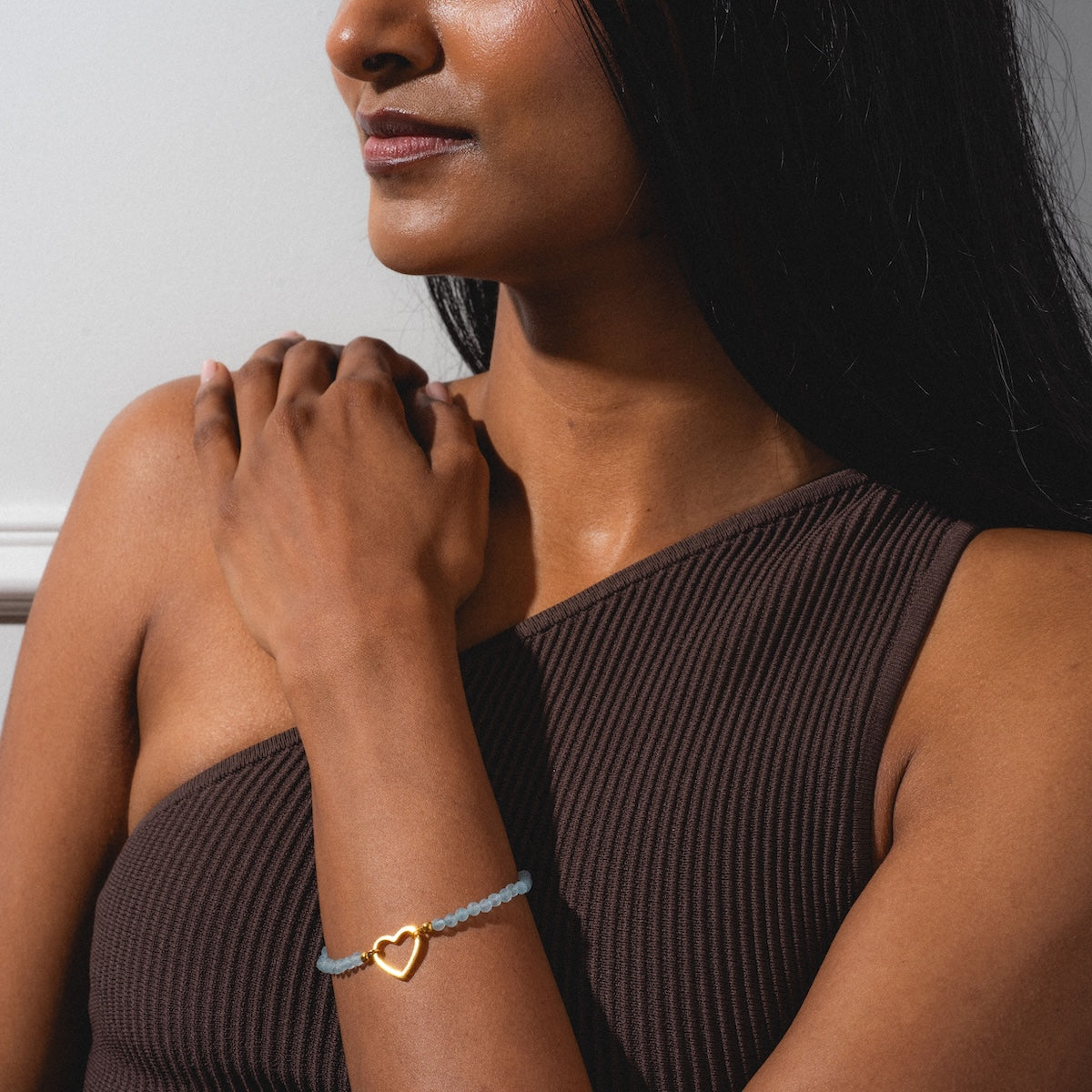 A woman in a ribbed, brown one-shoulder top rests her hand on her shoulder, showcasing the Aquamarine Beaded Heart Clasp Bracelet on her wrist against a softly lit, plain background.