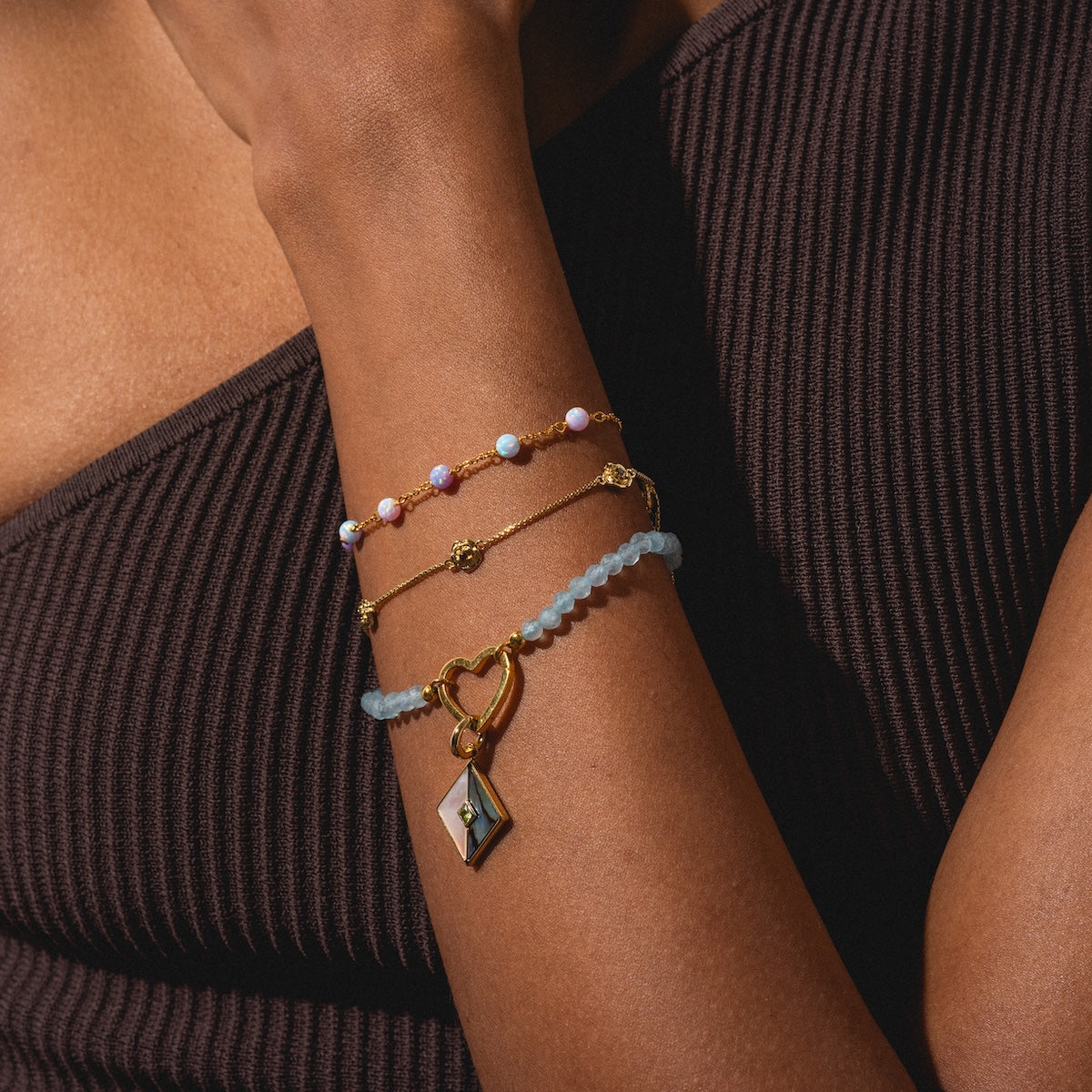 A person in a ribbed, dark sleeveless top wears three bracelets, including the Aquamarine Beaded Heart Clasp Bracelet with blue beads and a gold heart charm, plus gold and pink-blue-white beaded bracelets.