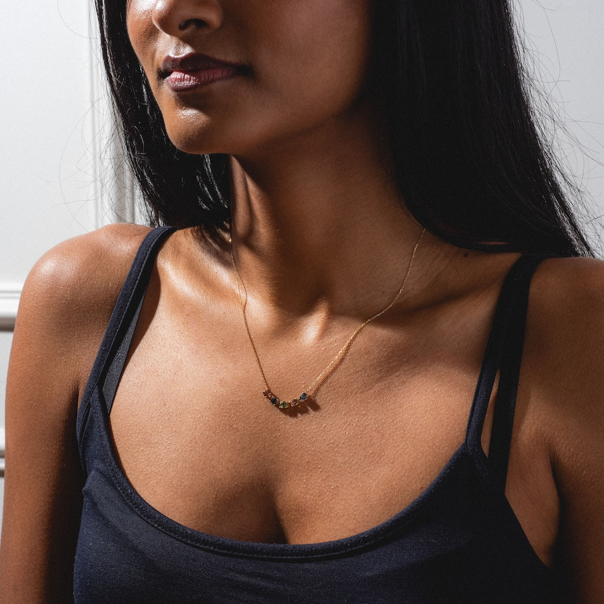 A woman shown from the shoulders up, against a plain background, wears a black tank top and the Royal Mile Necklace—a delicate gold necklace adorned with small colored stones.