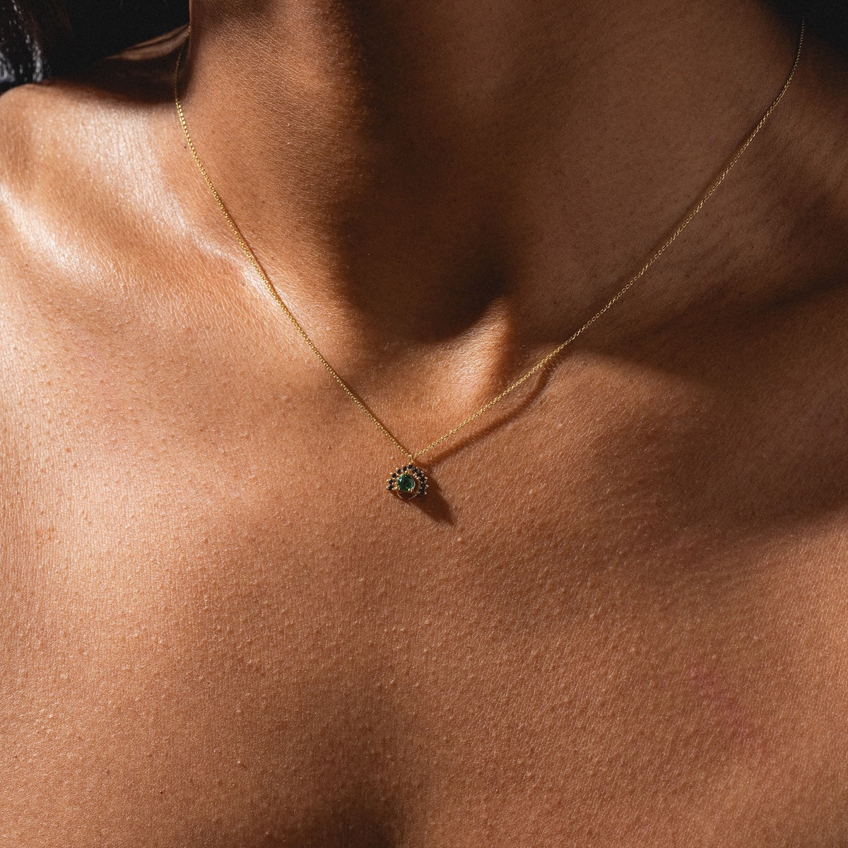 A close-up of a person’s neck and upper chest showcases the Tosia Black Diamond Necklace, featuring a delicate gold chain with a small, round black diamond pendant. The lighting highlights the wearer’s smooth skin and the elegant jewelry.