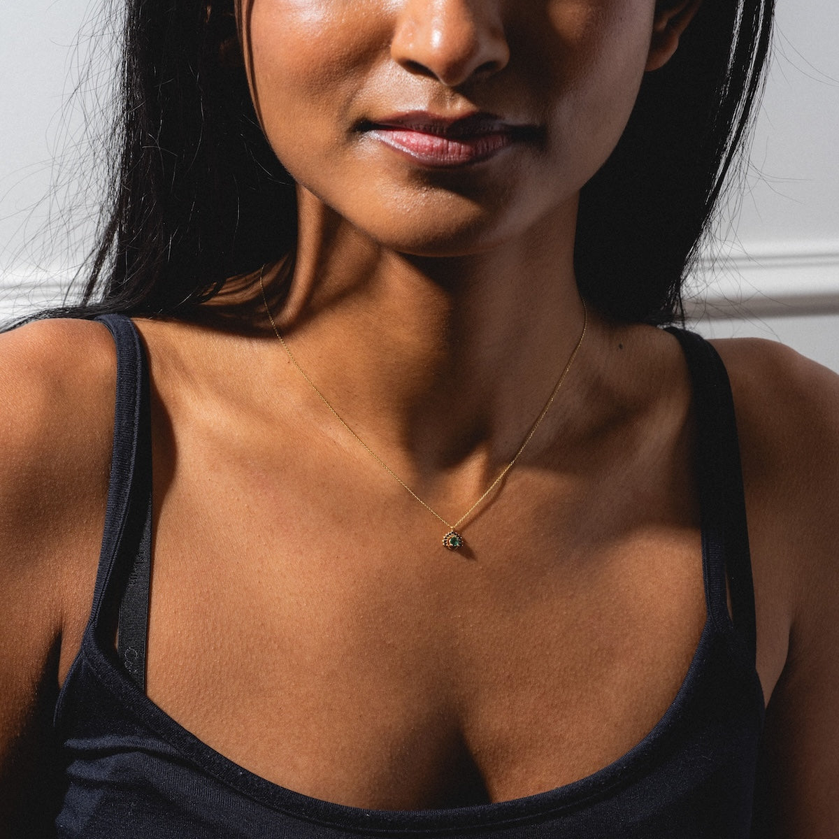 A woman wears a black tank top and the Tosia Black Diamond Necklace, featuring a delicate gold chain with a small green stone pendant, shown from the shoulders up against a neutral background.