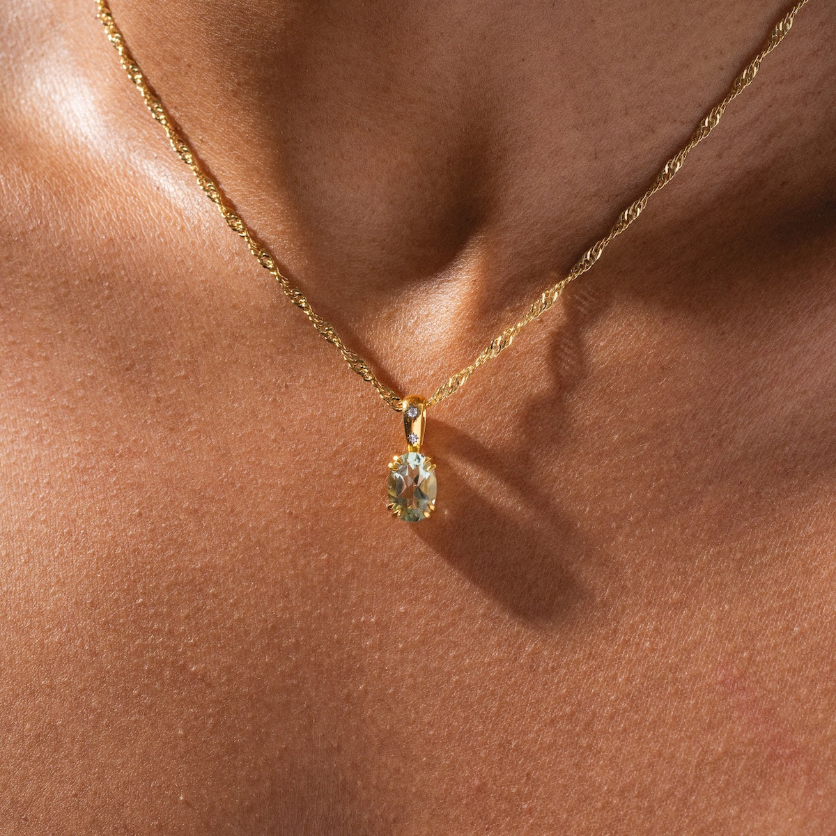 A close-up of a person’s neck wearing Charms by Carrie Elizabeth Jewelry—a twisted gold chain necklace with an oval-shaped gemstone pendant—shown against brown skin in natural lighting.
