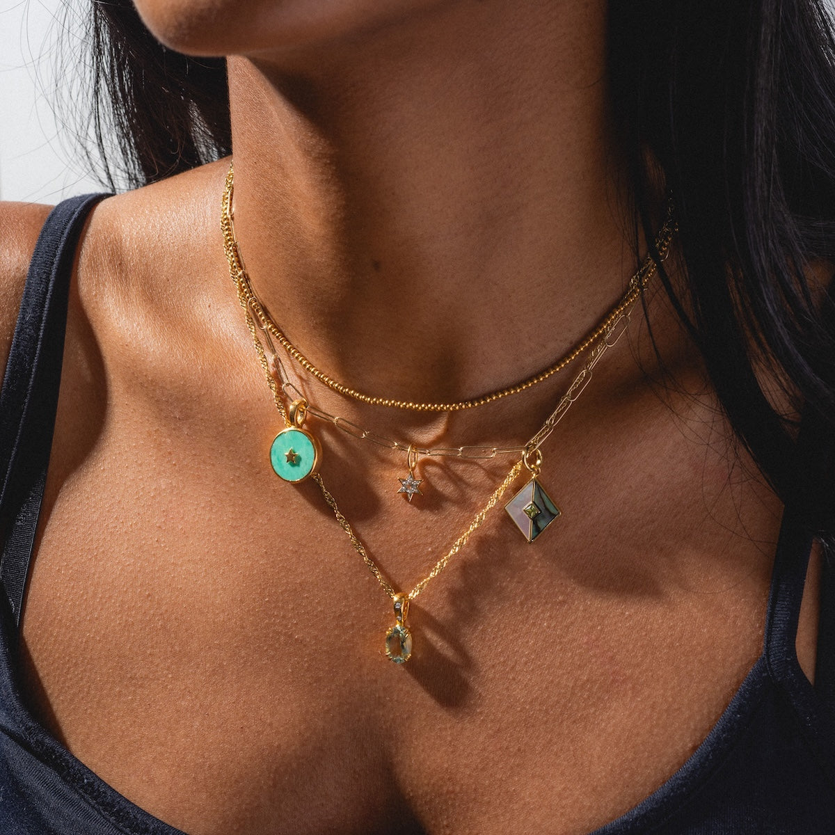 A woman in a black tank top showcases Charms by Carrie Elizabeth Jewelry: three layered gold necklaces featuring a turquoise circle, a small star, a rectangular gem, and a round green stone pendant.