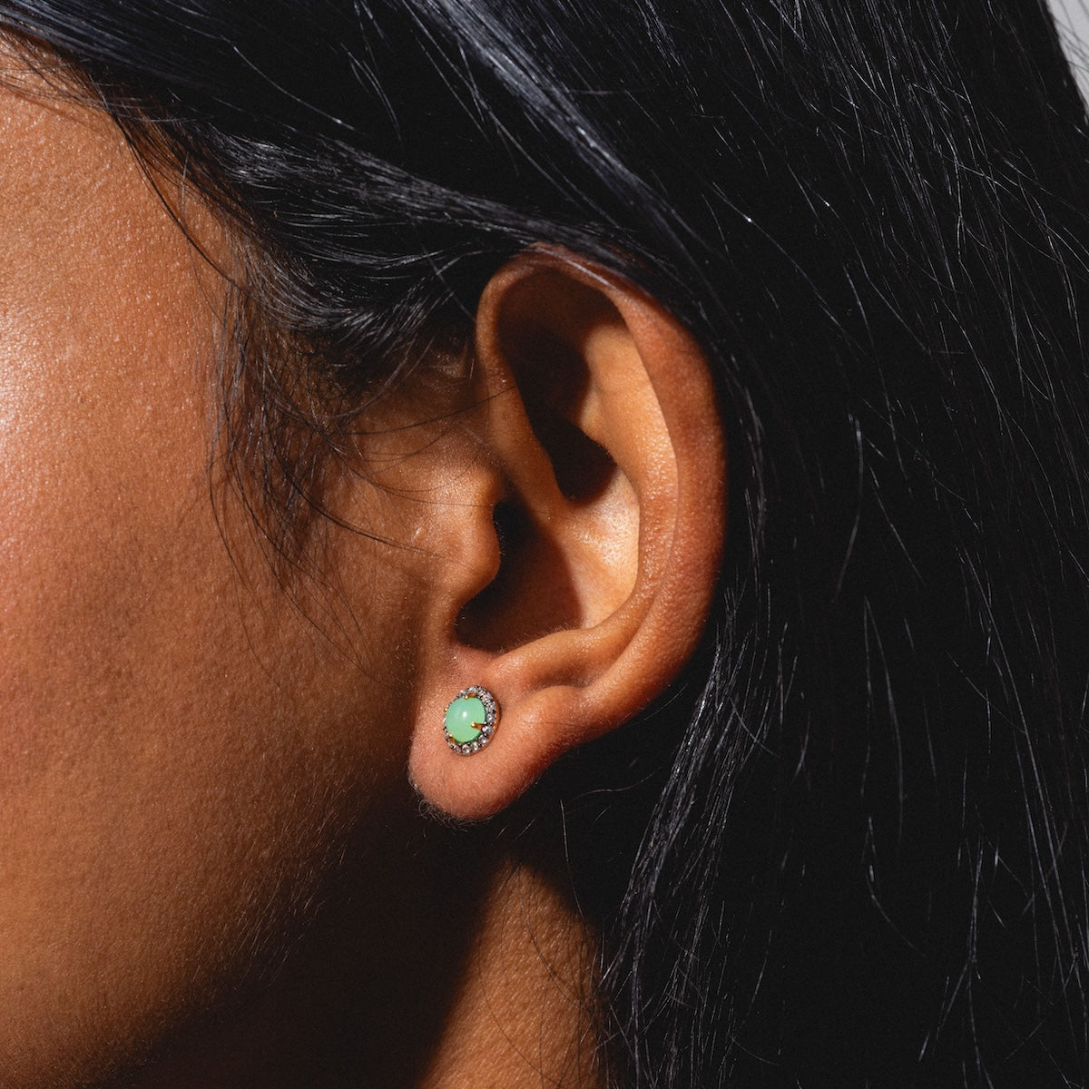 Close-up of a person with dark hair wearing Chrysoprase Pave Stud Earrings—small, round, light green studs encircled by tiny clear stones—against a neutral background.