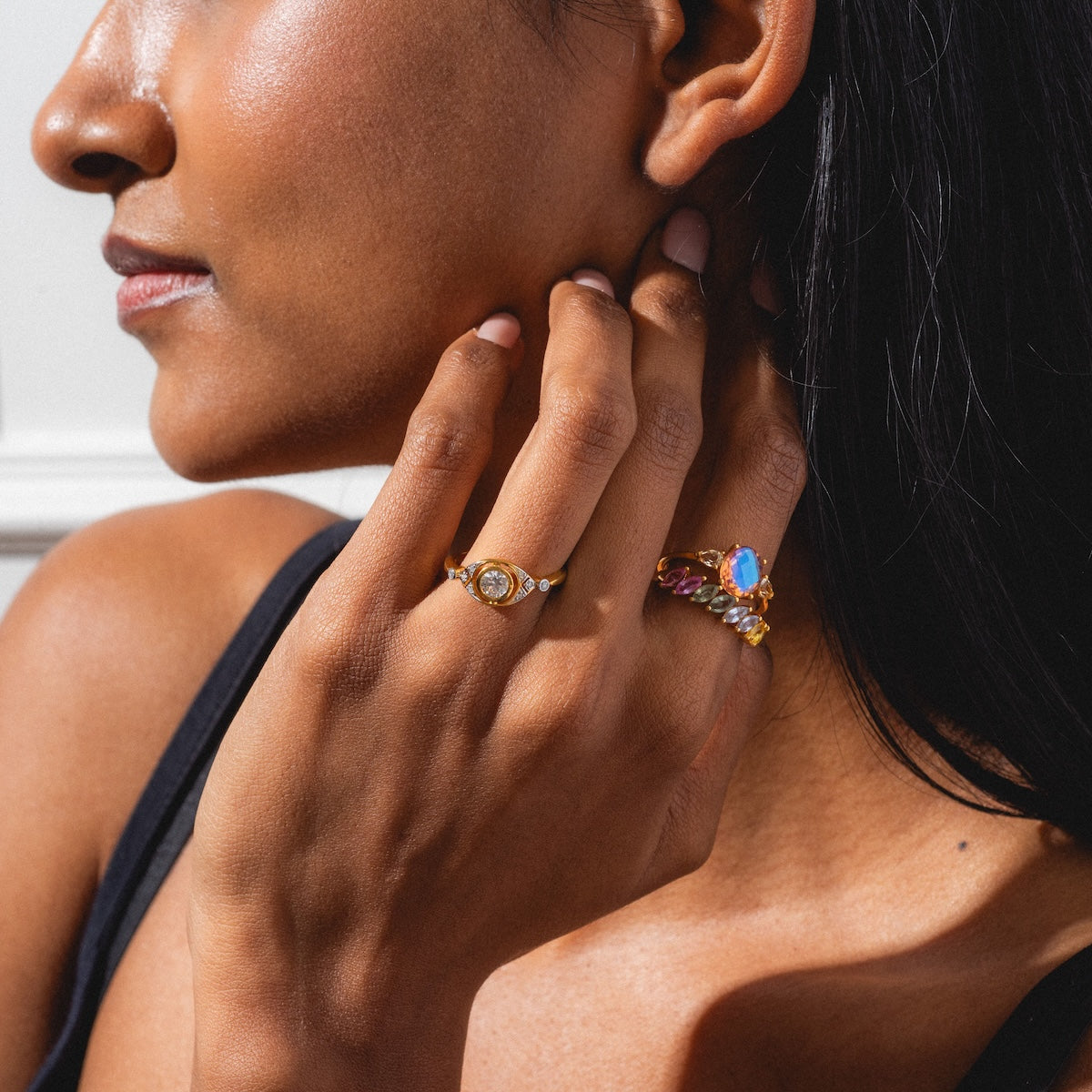 A woman with medium brown skin, straight dark hair, and a black top touches her neck. She wears two rings: a gold ring with a white stone and the Della Rainbow Sapphire Ring on another finger.