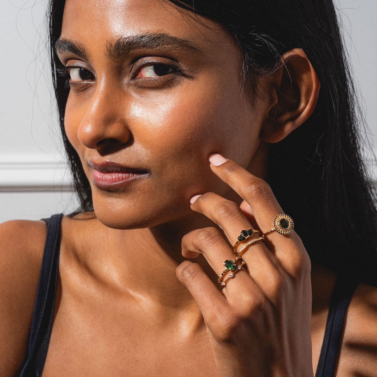 A woman with long dark hair and glowing skin wears a black top, smiling softly as she displays three Munro Rings—gold bands with green stones and sunburst designs—while holding her hand near her face.