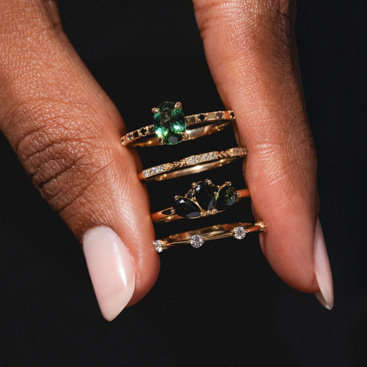 A manicured hand holds four Munro Rings—gold bands with green and clear gemstones in solitaire and cluster settings—against a dark background.