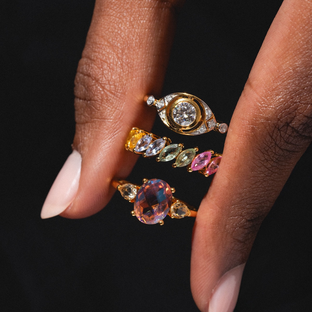 A close-up of a hand with light pink nails wearing the Della Rainbow Sapphire Ring, a gold ring featuring colorful sapphires in a unique design, against a dark background.