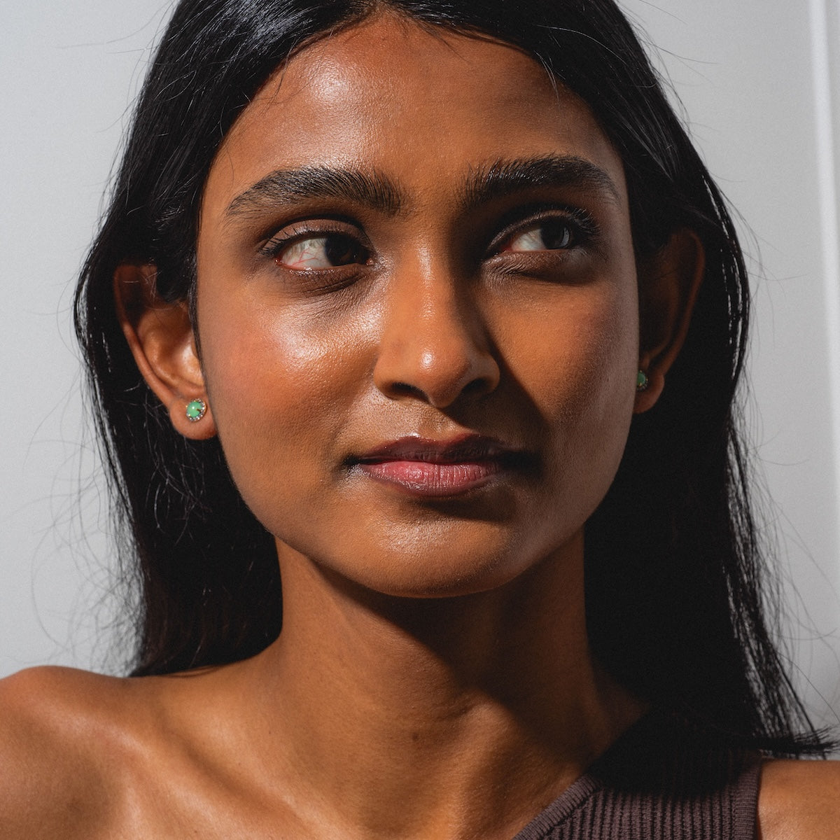 A woman with long dark hair and glowing skin gazes slightly left, wearing a one-shoulder brown top and Chrysoprase Pave Stud Earrings, her expression neutral under soft lighting.