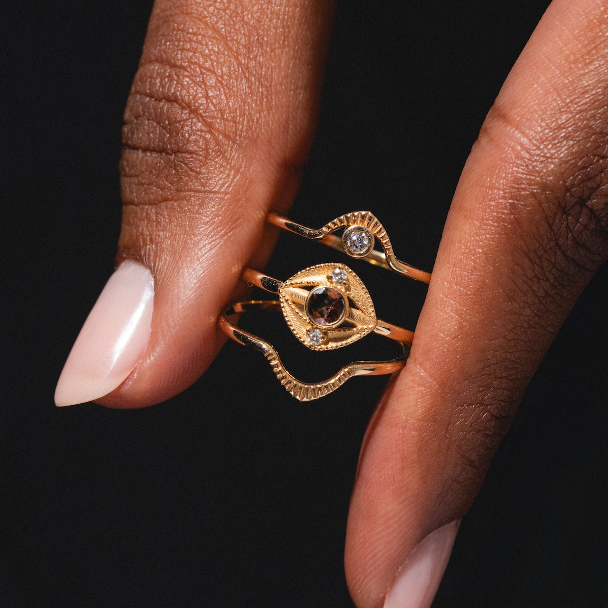 A hand with neatly manicured nails holds three Corrie Rings—delicate gold bands with intricate designs and small gemstones—set against a black background.
