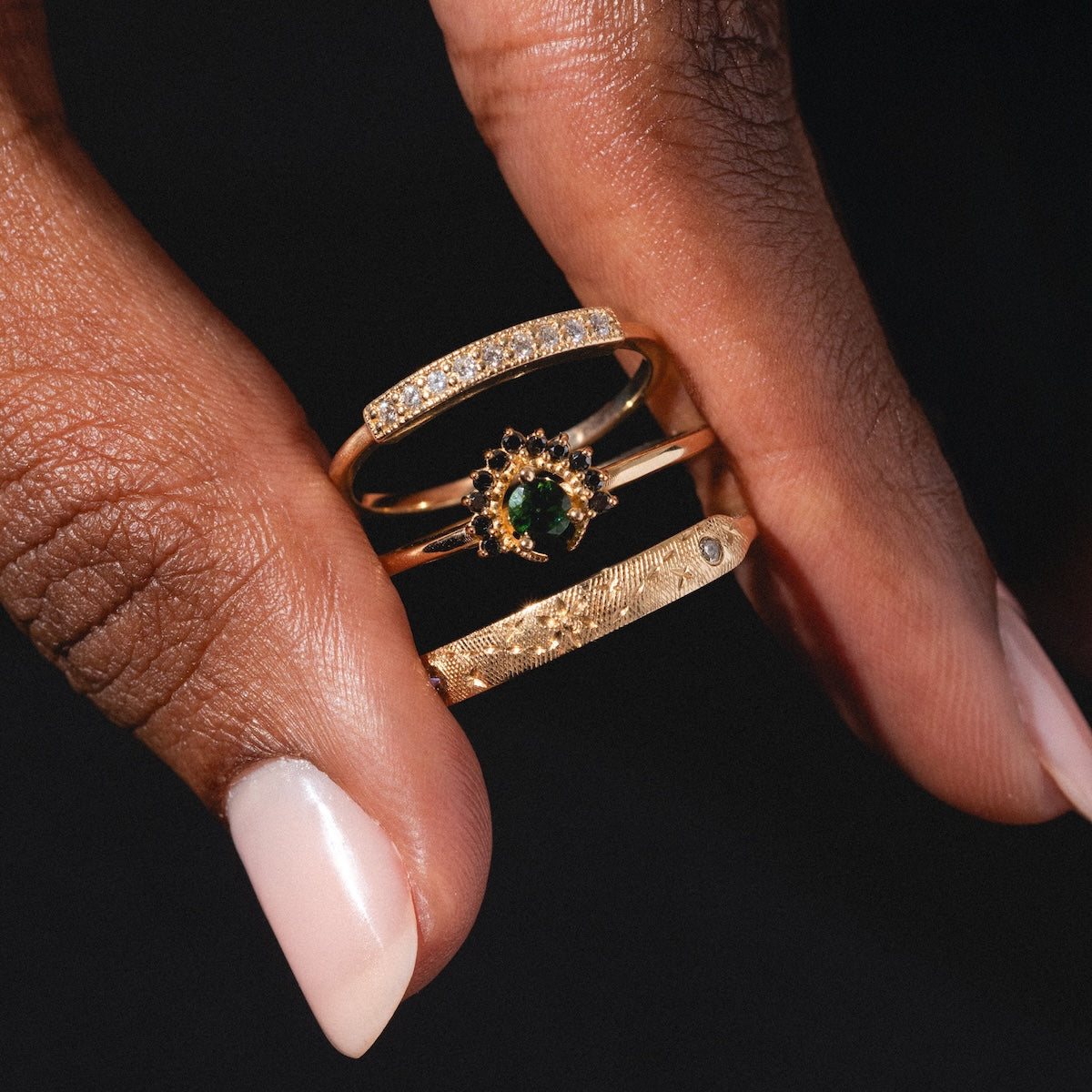 A hand with light pink manicured nails holds three stacked gold rings, including the Tosia Tourmaline Ring with an elegant center stone, alongside a diamond band and a textured gold ring.