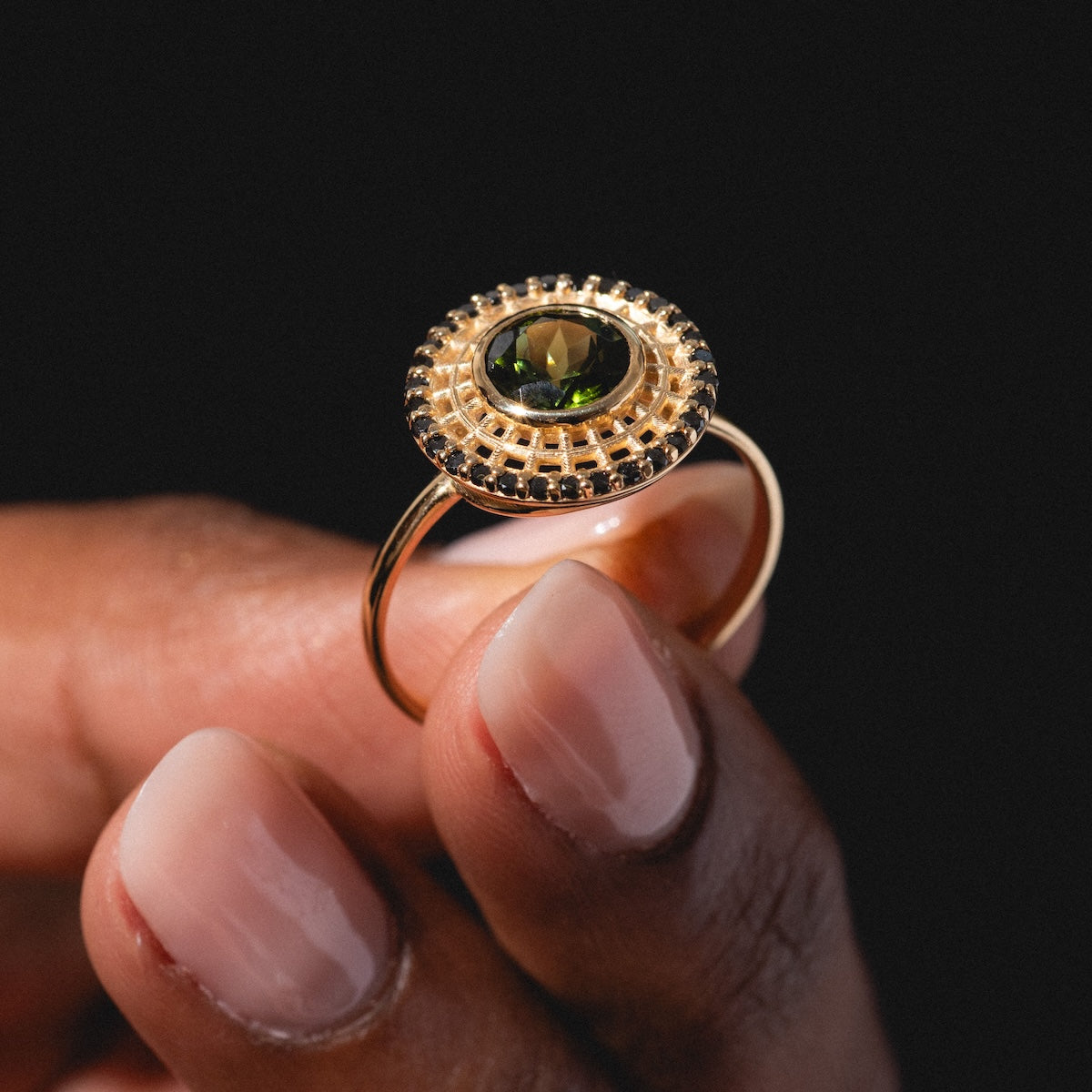 A hand with neatly manicured nails holds the Tantallon Black Diamond Ring, featuring a round black diamond in an ornate setting, shown against a black background.