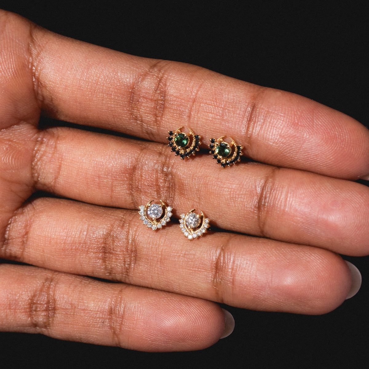 A close-up of a hand holding two pairs of Tosia Diamond Studs: one pair with green gemstones in gold settings, the other featuring clear stones encircled by small pearls or crystals, all set against a black background.