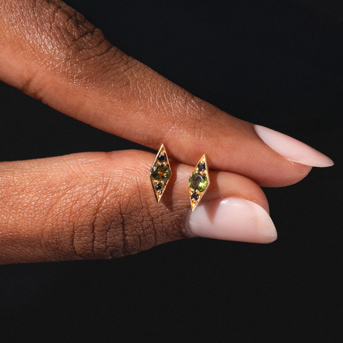 A hand with manicured nails holds the Shillay Studs—gold marquise-shaped earrings adorned with small green gemstones—against a dark background.