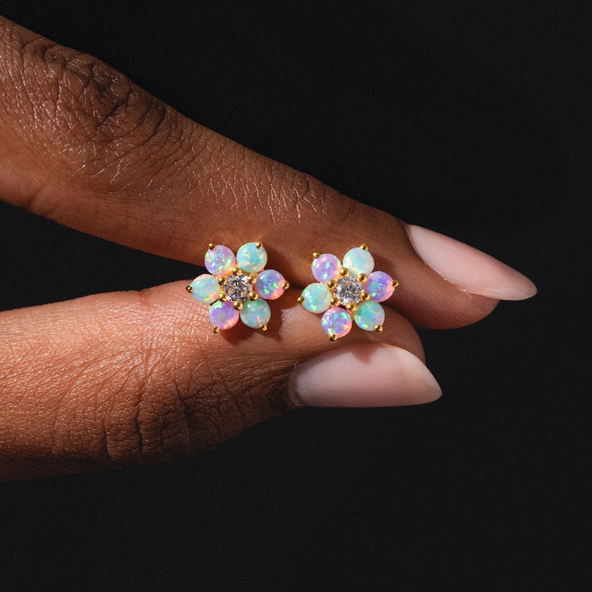 A hand with manicured nails holds the Tropical Opal Fleur Studs—gold flower-shaped earrings with six iridescent opal petals and a cluster of clear gems at the center—against a black background.