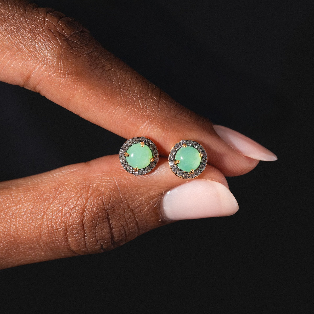 A hand with manicured nails holds Chrysoprase Pave Stud Earrings—round green gemstones encircled by a halo of small white stones—against a dark background.