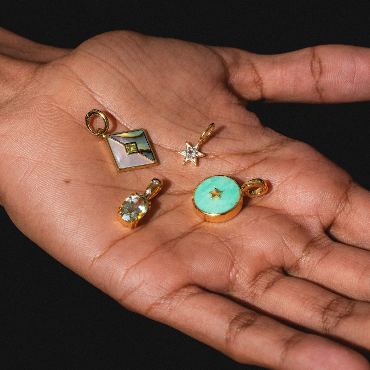 A hand displays four Charms by Carrie Elizabeth Jewelry: a square mother-of-pearl, a gem star, an oval gemstone, and a round turquoise pendant with a gold star cutout.