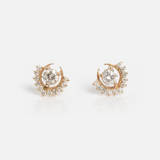 The Tosia Diamond Studs are gold earrings with a central round diamond surrounded by smaller sparkling stones in a crescent shape, featuring an elegant, intricate design and displayed on a white background.