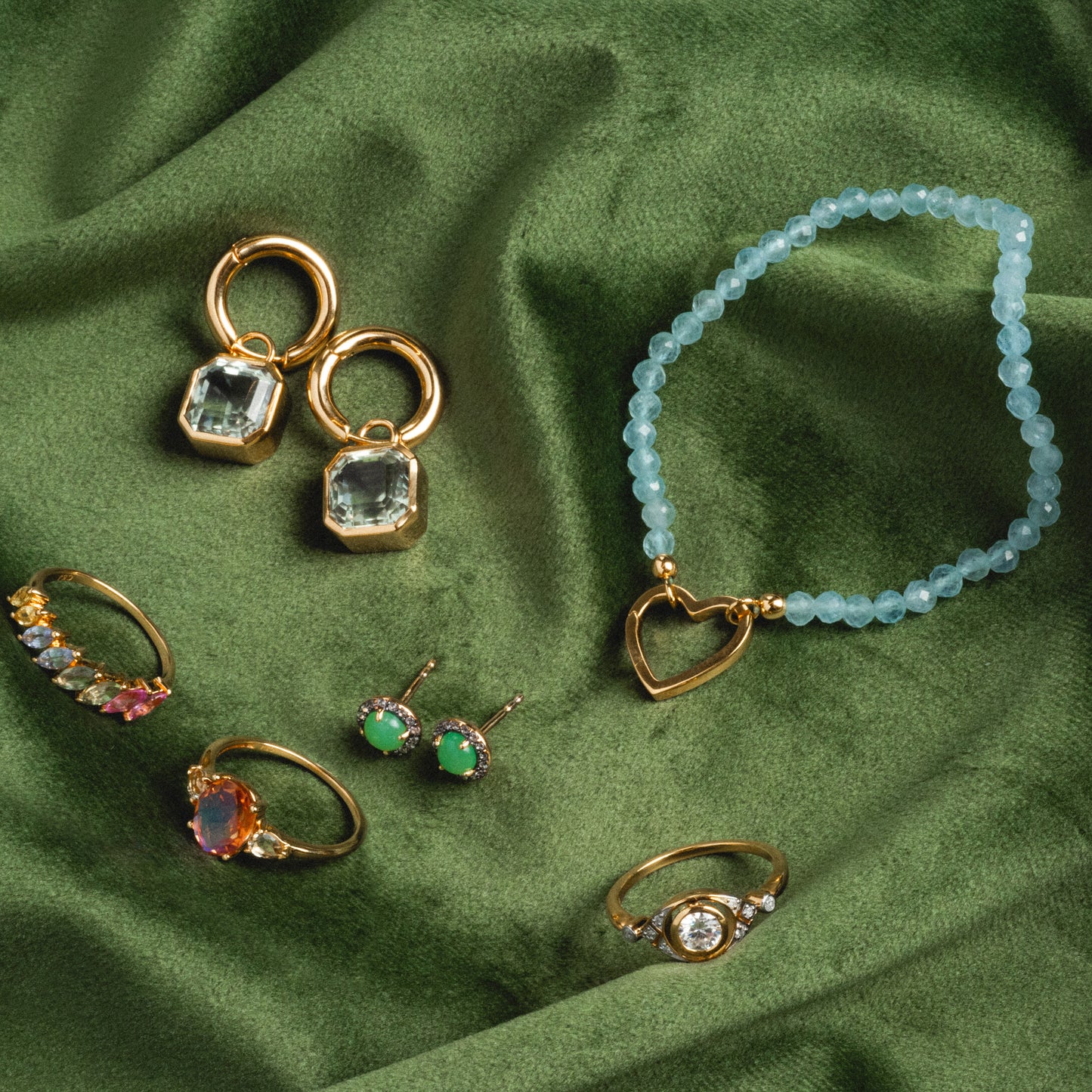 The Vienna Moissanite and Diamond Ring, along with gold earrings and a green beaded necklace with a heart pendant, are displayed on green velvet. The vintage-inspired ring showcases sparkling gemstones and intricate detailing.