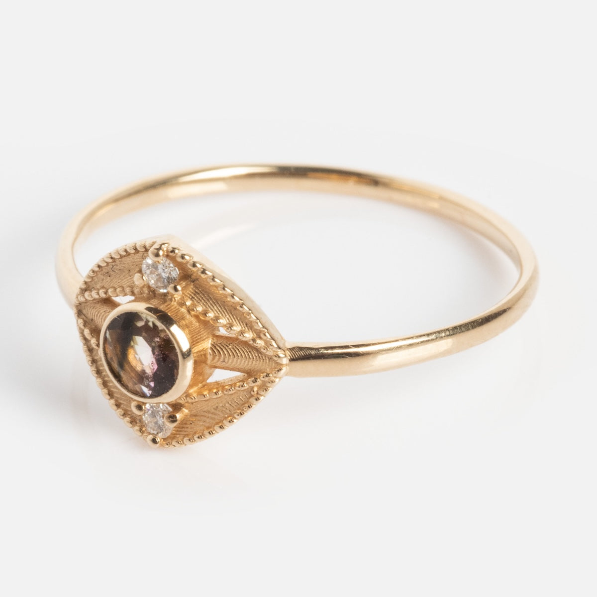 The Staffa White Diamond Ring is a delicate gold ring with an oval center gemstone, bordered by intricate detailing and three small clear stones on a thin gold band.