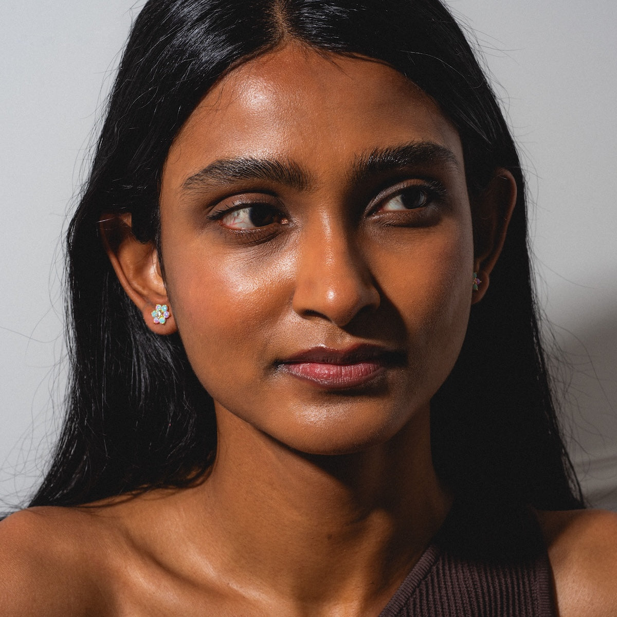 A woman with long dark hair and radiant skin wears the Tropical Opal Fleur Studs—flower-shaped earrings—paired with a brown, one-shoulder top. Soft lighting highlights her features against a simple background as she gazes to the side.